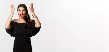Moda ve güzellik. Siyah elbiseli kızgın bir kadın, deli gibi bağırıyor ve el sıkışıyor, kamera karşısında suratını ekşitiyor, beyaz arka planda duruyor..