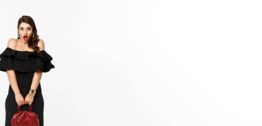 Güzellik ve moda konsepti. Uzun boylu, somurtan, şaşırmış görünen, el çantası tutan, topuklu ve siyah elbise giyen, beyaz arka planlı bir kadın..