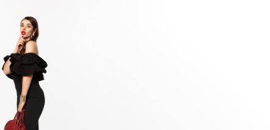 Güzellik ve moda konsepti. Siyah zarif elbiseli, yüksek topuklu ayakkabılı ve çantalı, uzun boylu, şehvetli genç bir kadın. Dudaklarına dokunuyor, kameraya bakıyor, beyaz arka plan..