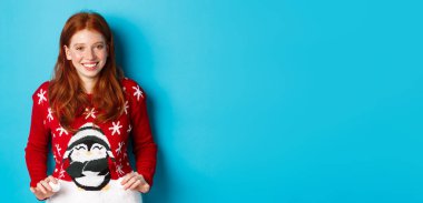 Kış tatili ve Noel arifesi konsepti. Şirin kızıl saçlı, sevimli kazak giyen ve gülümseyen, yeni yılı kutlayan, mavi arka planda duran..