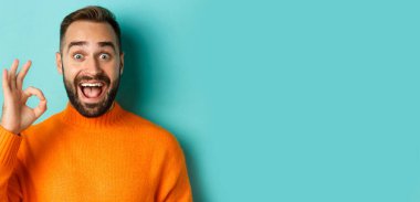 Etkilenmiş yakışıklı bir adam iyi bir işaret veriyor, iyi bir şey öneriyor, iyi bir jestle övüyor, açık mavi arka planda duruyor..