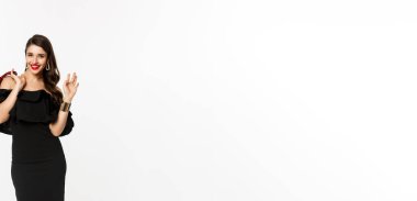 Güzellik ve moda konsepti. Cazibeli, şık, kendine güvenen siyah elbiseli bir kadın. Çantasını tutuyor ve öne doğru yürüyor. El sallayarak merhaba diyor..