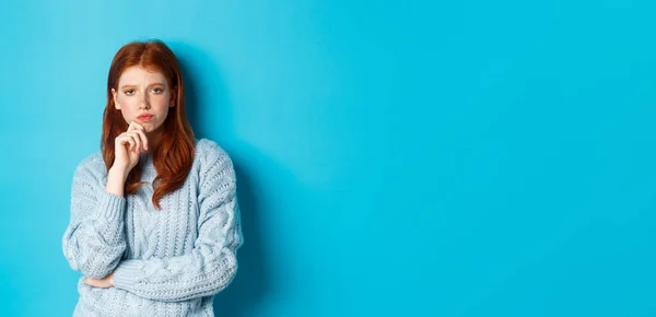 Thoughtful teenage redhead girl staring at camera, pondering ideas, making decision with serious face, standing over blue background.