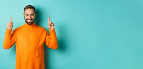 Handsome bearded guy in orange sweater showing up promo, pointing at top and smiling, standing over turquoise background.