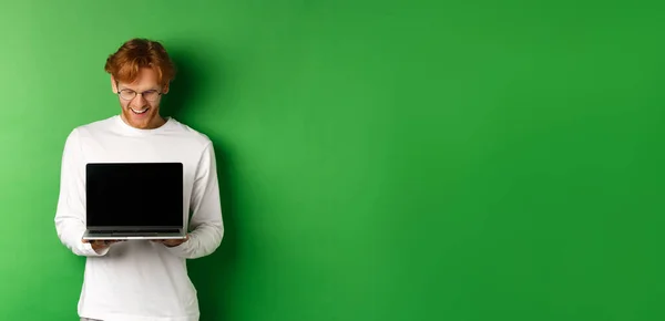 Happy young man with red hair, showing blank laptop screen and looking pleased, smiling while staring at computer, green background.