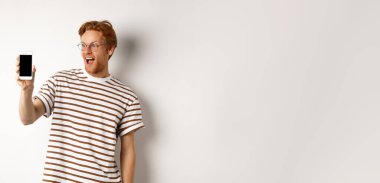 Teknoloji ve e-ticaret kavramı. Gözlüklü mutlu genç adam boş akıllı telefon ekranı gösteriyor. Şaşırmış görünüyor. Beyaz arka planda duruyor..