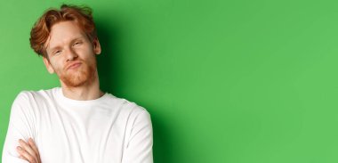 Duygular ve moda konsepti. Kızıl saçlı, sakallı, büzüşmüş dudakları olan, kameraya bakan, yeşil arka planda duran şüpheci genç adamın yakın çekimi..