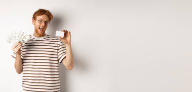 Alışveriş ve finans kavramı. Gözlüklü genç kızıl adam plastik kredi kartını seçiyor, dolar gösteriyor ve gülümsüyor, beyaz arka planda duruyor..