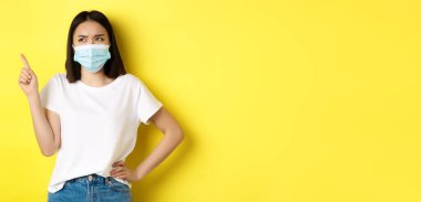 Covid, sağlık ve salgın hastalık konsepti. Tıbbi maskeli ve beyaz tişörtlü Asyalı kadın sol üst köşedeki logoyu işaret ediyor..