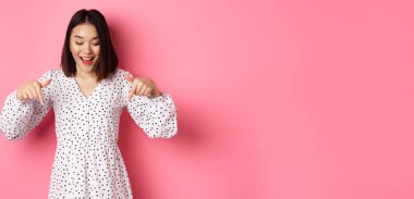 Romantik Asyalı kadın dikizliyor, parmakları dipte, ürün indirimine bakıyor mutlu bir gülümsemeyle, pembe arka planda duruyor..