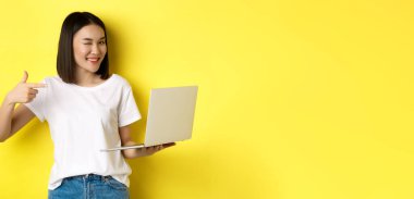 E-ticaret ve alışveriş konsepti. Güzel görünümlü Asyalı kız kameraya göz kırpıyor, gülümsüyor ve bir dizüstünü işaret ediyor, bir şey gösteriyor, sarı arka planda duruyor..