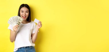 Kısa siyah saçlı, beyaz tişörtlü, parayı dolar ve kredi kartlarıyla gösteren, sarı arka planda duran güzel Asyalı kadın..