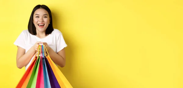 Happy asian female shopper smiling and holding colorful shopping bags, standing against yellow background.
