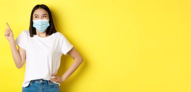 Covid, sağlık ve salgın hastalık konsepti. Tıbbi maskeli ve beyaz tişörtlü Asyalı kadın sol üst köşedeki logoyu işaret ediyor..