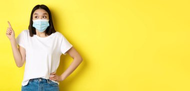 Covid, sağlık ve salgın hastalık konsepti. Tıbbi maskeli ve beyaz tişörtlü Asyalı kadın sol üst köşedeki logoyu işaret ediyor..