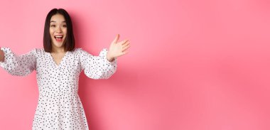 Mutlu Asyalı kız ellerini kameraya uzatıyor, sarılmaya uzanıyor, gülümsüyor ve bir şeyi tutmak istiyor, pembe arka planda duruyor..