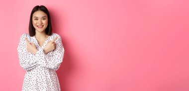 Romantik elbiseli güzel Asyalı kız, iki farklı alışveriş çeşidini gösteriyor, kameraya gülümsüyor, pembe arka planda duruyor..