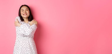 Güzel Asyalı bir kadın romantik hissediyor, kendine sarılıyor, gözlerini kapatıyor ve rüya gibi yüzüyle gülümsüyor, pembe arka planda mutlu bir şekilde duruyor..