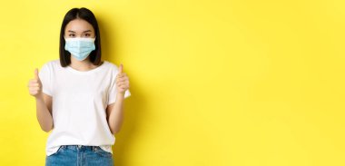Covid-19, karantina ve sosyal uzaklık kavramı. Tıbbi maskeli ve beyaz tişörtlü neşeli Asyalı kadın onaylanmış başparmaklarını gösteriyor, iyi bir anlaşma, sarı arka plan.