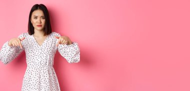 Hayal kırıklığına uğramış Asyalı kadın manken tanıtım teklifini işaret ediyor. Kaşlarını çatıyor ve kameraya bakıyor. Pembe arka planda dikiliyor..