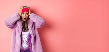 Şok olmuş ve paniklemiş Asyalı yaşlı bir kadın, mor sahte kürk giyiyor, el ele tutuşuyor ve panikliyor, pembe arka plana karşı tedirgin duruyor..