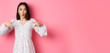 Etkilenmiş Asyalı kadın parmakları aşağıda, şuraya bak, indirimlere ve satışlara bak, pembe arka planda şaşkın şaşkın duruyor..