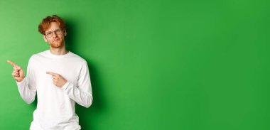 Gözlüklü, solu işaret eden, kameraya tereddütlü ve kararsız bakan, yeşil arka planda duran şüpheli kızıl saçlı adam..