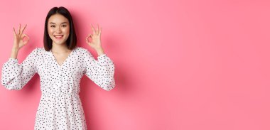Hoş, genç Asyalı bir kadın iyi bir işaret veriyor, övgüler yağdırıyor ve onaylıyor, tatmin olmuş görünüyor, pembe arka plana karşı duruyor..