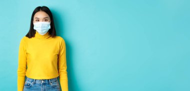 Covid-19, sosyal uzaklık ve salgın konsepti. Tıbbi maskeli genç Asyalı kadın, kameraya bakıyor ve sarı kazaklı mavi arka planda duruyor..