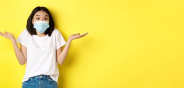 Covid-19, karantina ve sosyal uzaklık kavramı. Tıbbi maskeli cahil Asyalı kadın omuz silkiyor, ellerini yana açıyor hiçbir şeyden haberi yok, sarı arka planda..