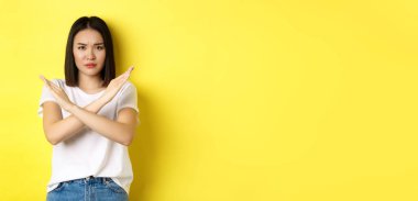 Ciddi ve iddialı Koreli kadın dur işareti yapıyor, kaşlarını çatıyor ve hayır diyor, eylemi yasaklıyor, kötü bir şeyi onaylamıyor, sarı arka planda duruyor..