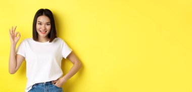 Kendine güvenen Asyalı kadın gülümsüyor ve tamam işareti gösteriyor. Onaylıyor ve iyi bir teklif sunuyor. Sarı arka planda beyaz tişörtle duruyor..