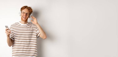 Teknoloji konsepti. Kızıl saçlı ve sakallı genç adam kulaklıkla müzik dinliyor ve akıllı telefon kullanıyor, kameraya gülümsüyor, beyaz arka plan.