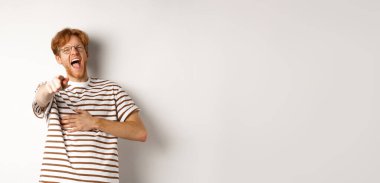 Kızıl saçlı ve sakallı genç adam kamerayı işaret ediyor ve gülüyor, komik biriyle dalga geçiyor, beyaz arka planda duruyor..