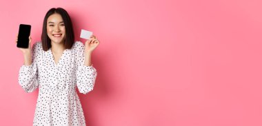Akıllı telefon ekranı ve kredi kartı gösteren neşeli Koreli kadın, internet siparişini ödüyor, online alışveriş uygulaması gösteriyor, pembe arka planda duruyor..