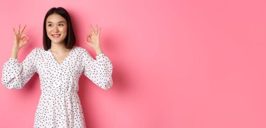 Hoş, genç Asyalı bir kadın iyi bir işaret veriyor, övgüler yağdırıyor ve onaylıyor, tatmin olmuş görünüyor, pembe arka plana karşı duruyor..