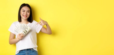 Genç Asyalı kadın gülümsüyor, ödül parasını gösteriyor, dolarları işaret ediyor, sarı arka planda duruyor..