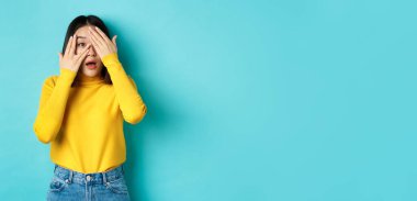 İlgi çekici Asyalı kız ilginç bir şeye parmak ucuyla bakıyor, kameraya bakıyor, mavi arka planda duruyor..