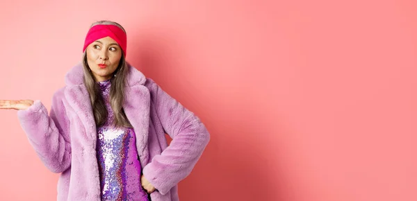 Fashion and shopping concept. Stylish asian senior woman in purple winter outfit demonstrating product on hand. Cool old lady in trendy clothes holding something on palm, pink background.
