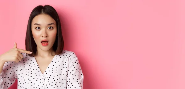 Image of confused asian woman pointing at herself, drop jaw and staring questioned at camera, standing over pink background.