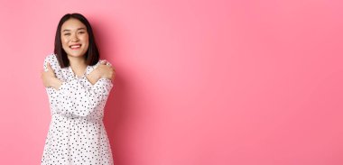 Güzel Asyalı kız kendi vücudunu kucaklıyor, kendini kucaklıyor ve mutlu bir yüzle gülümsüyor, kaygısız bir şekilde kameraya bakıyor, pembe arka planda duruyor..