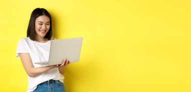 Dizüstü bilgisayarda çalışan sevimli Asyalı kız öğrenci, ekranda okuyor ve gülümsüyor, sarı arka plan..