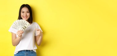 Genç Asyalı kadın gülümsüyor, ödül parasını gösteriyor, dolarları işaret ediyor, sarı arka planda duruyor..