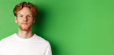 Kızıl saçlı, sakallı, yeşil arka planda duran ve kameraya bakan genç adamın yakın çekimi..