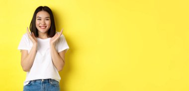 Güzellik ve moda konsepti. Heyecanlı Asyalı kadın el çırpıyor ve kameraya mutlu bir şekilde gülümsüyor, beyaz tişörtle sarı arka planda duruyor..