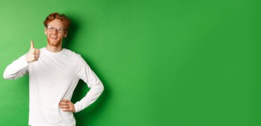 Sakallı, kızıl saçlı, gözlüklü, onaylanmış ve gülümseyen, yeşil arka planda duran neşeli genç adam..