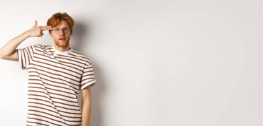 Sinirli genç kızıl saçlı adam parmak tabancasını kafasının yakınında yapıyor, kameraya bakıyor ve rahatsız ediyor, beyaz arka plan..