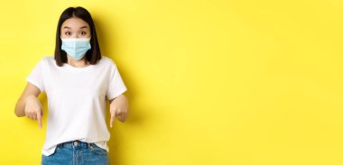 Covid-19, salgın hastalık ve sosyalleşme konsepti. Tıbbi maskeli şaşırmış Asyalı kadın, reklam veriyor, sağa bakıyor ve gülümsüyor, sarı arka plan.