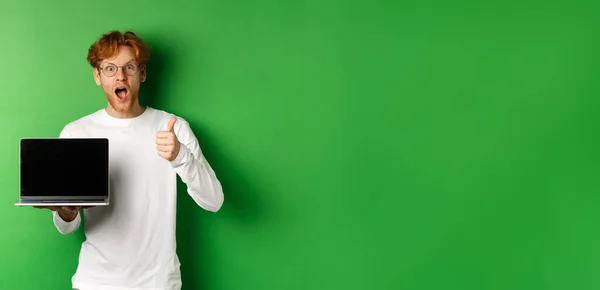 Impressed caucasian man with red hair and beard, showing blank laptop screen and thumb-up, looking amazed at camera, praising something online, green background.