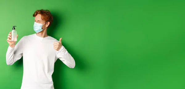 Covid-19, virus and social distancing concept. Handsome young man with red hair, wearing medical mask, looking at antiseptic and showing thumb-up, green background.